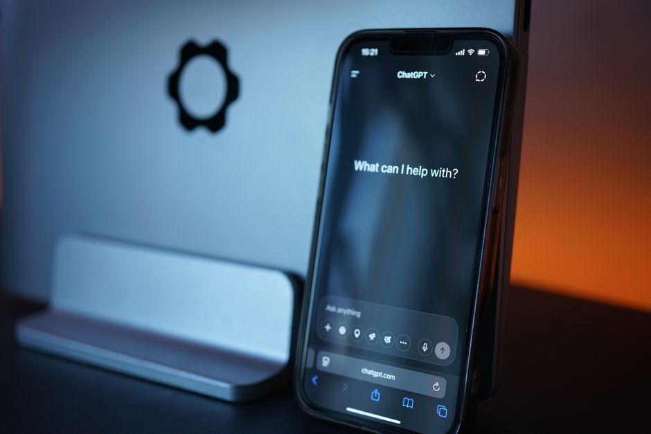 A smartphone on a desk showing an open AI chat app asking "What can I help with?", representing asking a chatbot who unfollowed you on Instagram