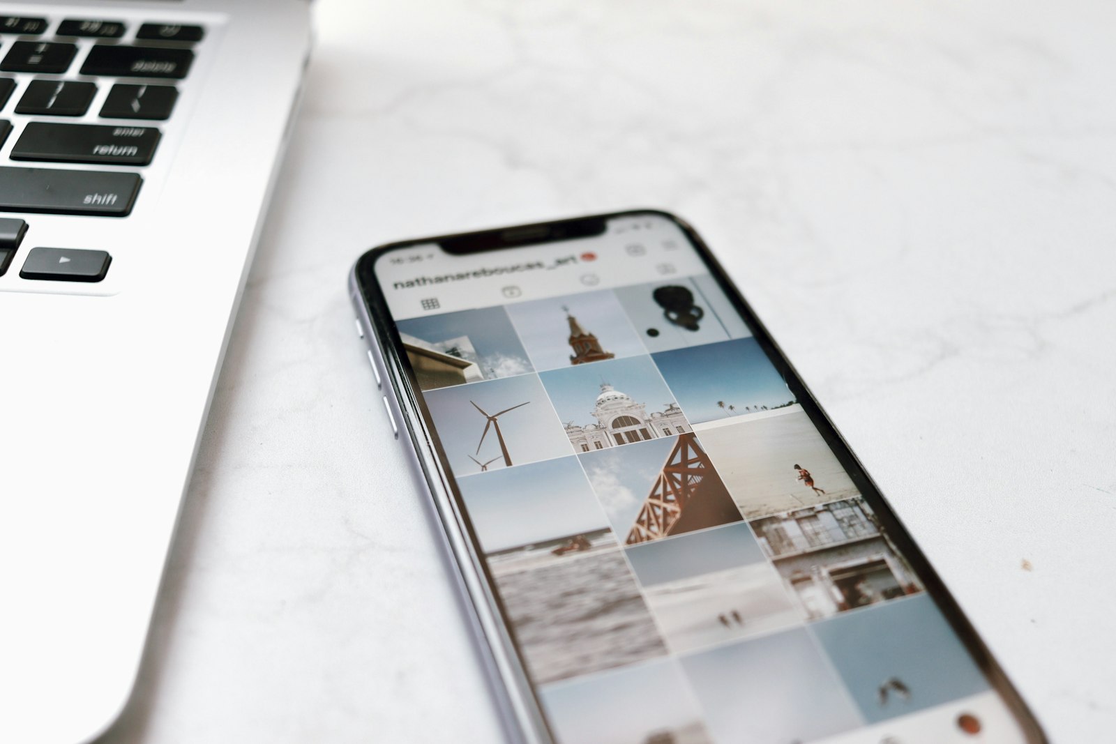 A phone on a marble desk beside a laptop, showing an Instagram profile feed